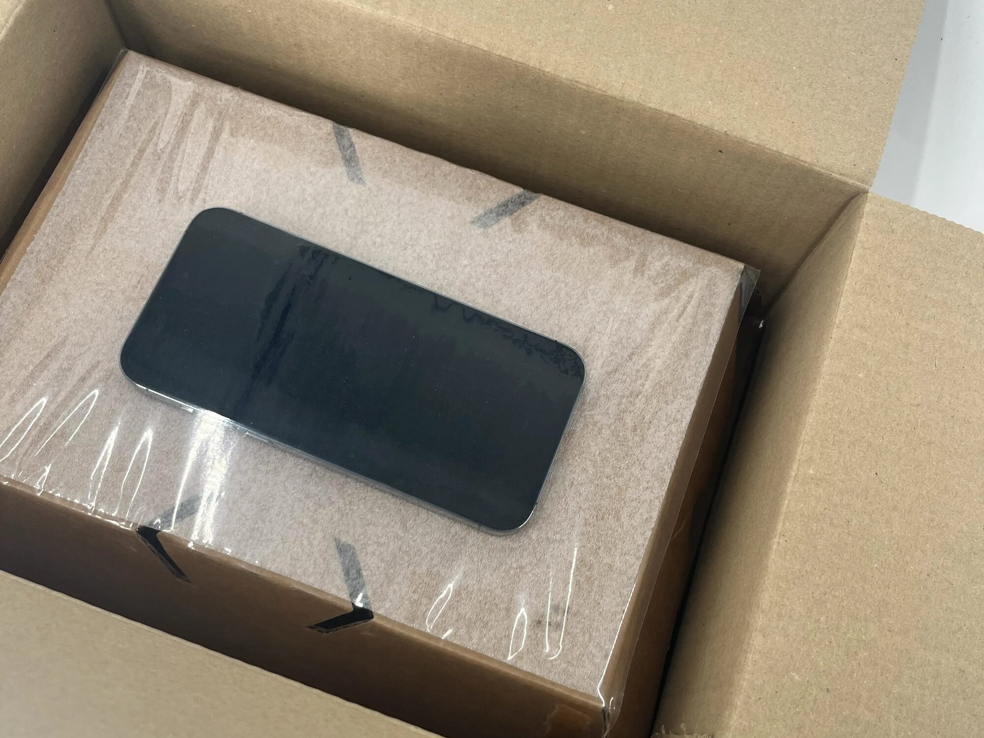 A smartphone held against a cardboard support by clear retention film inside an open corrugated box.