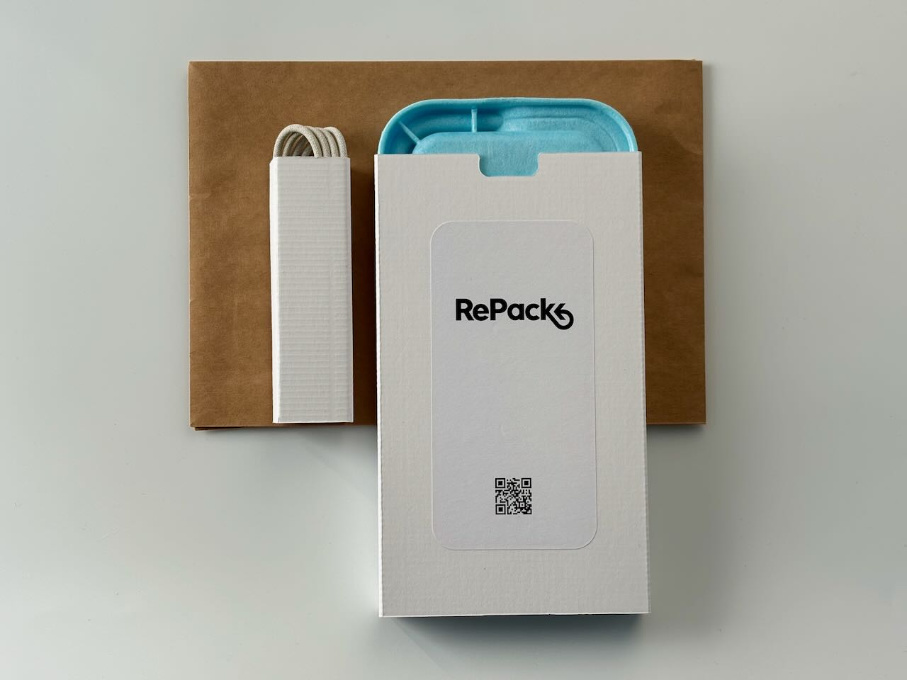A white RePack Vie sleeve printed with the RePack logo and a QR code, a blue tray edge visible at its opening, beside a white corrugated holder with a coiled cable, both resting on a kraft paper envelope.